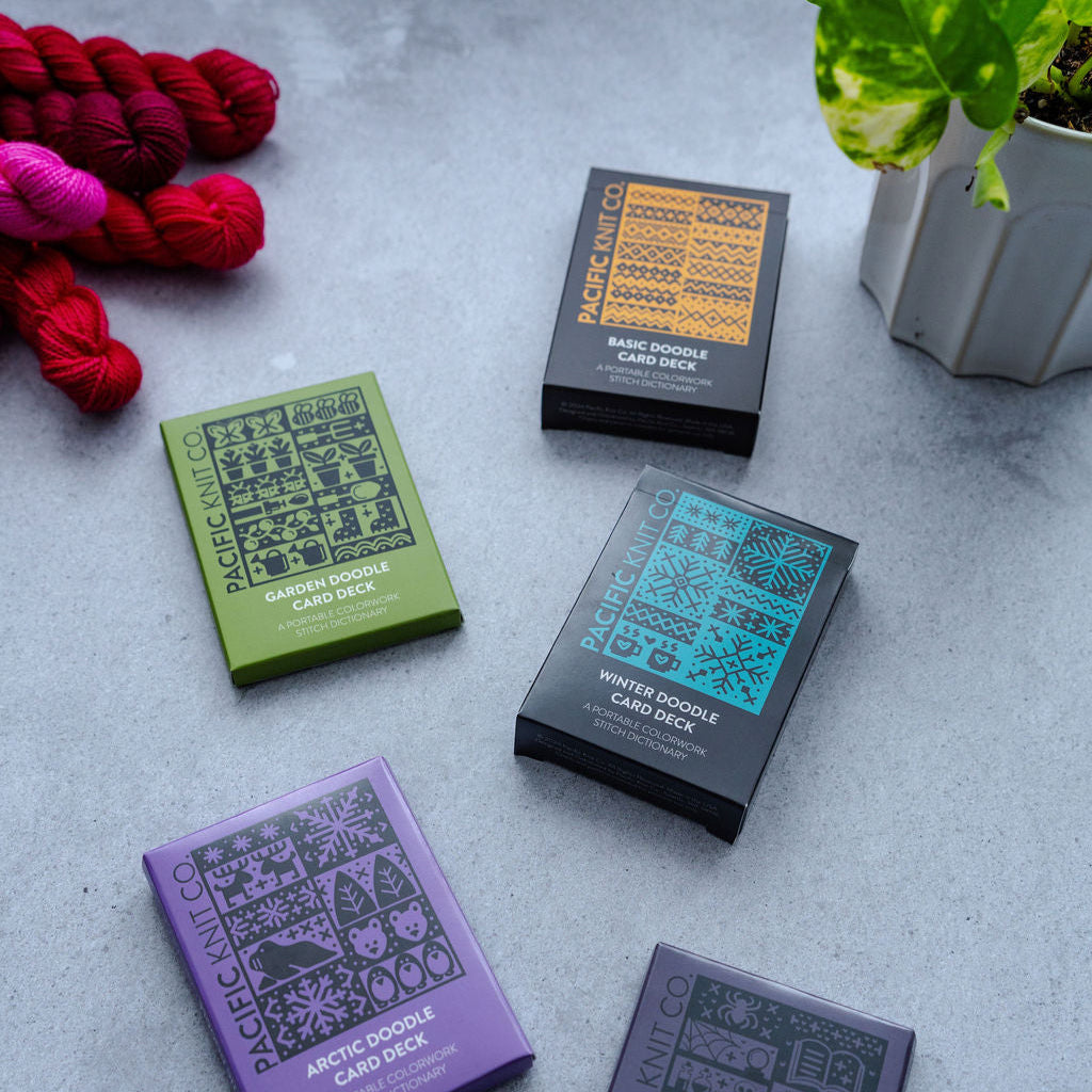 Card decks with colorful packaging on a grey surface, surrounded by yarn and a plant.